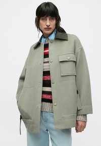 Svetlo zelena jakna iz volnenega mešanega materiala s kontrastnim temnim ovratnikom, zapiranjem na pritisne gumbe, sprednjim žepom in stranskimi razporki. Nosa se čez črtasti pulover.