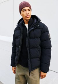 Chaqueta de plumas negra con capucha, cierre de cremallera y puños acanalados. Llevada sobre un suéter oscuro, complementada con pantalones verde oliva.
