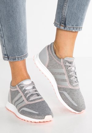 Fødder iført grå og lyserøde snørestøvler kombineret med lyseblå op rullede jeans, stående på en hvid overflade.