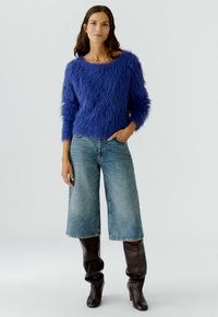Frau in blauer, flauschiger Sweater, knielangen Denim-Culottes und dunkelbraunen, weichen Stiefeln mit Absatz, steht mit einer Hand in der Tasche.