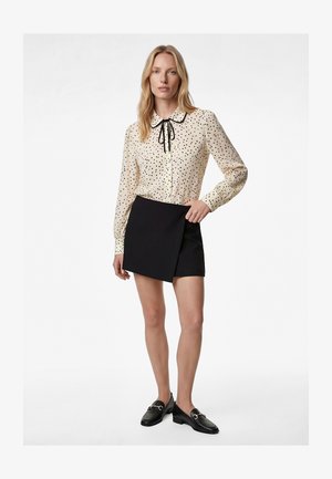 Vrouw draagt een crème blouse met zwarte polkadots en een strik, gecombineerd met een zwarte asymmetrische minirok en zwarte loafers.