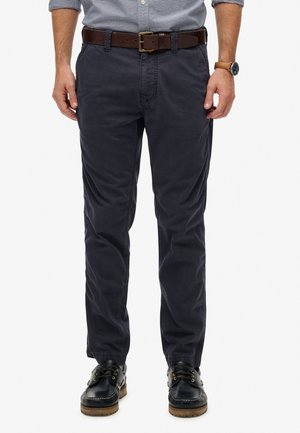 Mężczyzna w granatowych chino, brązowym skórzanym pasku, czarnych mokasynach i koszuli w kratę z podwiniętymi rękawami, stojący na białym tle.