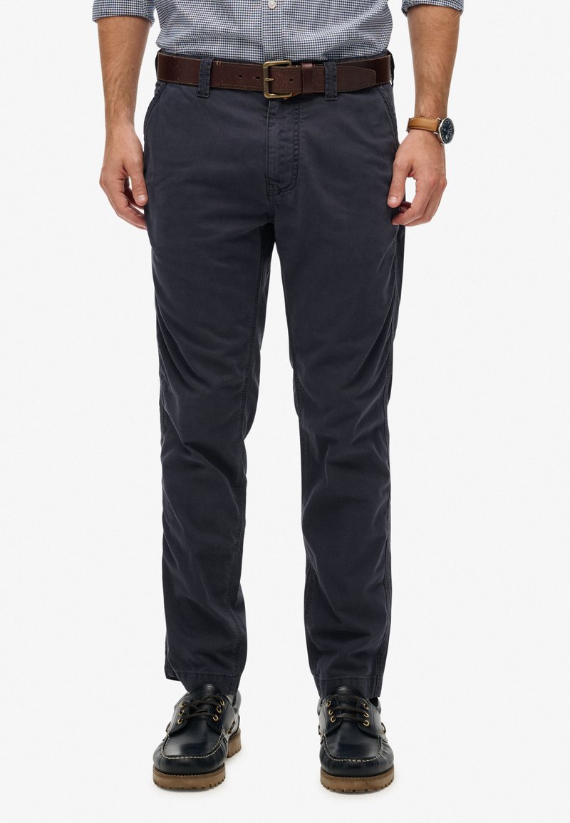 Uomo con chino blu navy, cintura in pelle marrone, scarpe da barca nere e camicia a quadri con maniche arrotolate, in piedi su sfondo bianco.