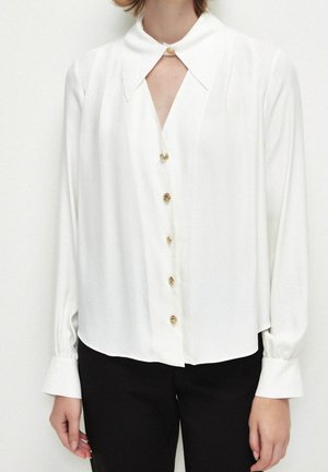 Witte blouse met lange mouwen, gouden knopen en een puntkraag, gecombineerd met zwarte broek bij een persoon tegen een effen achtergrond.