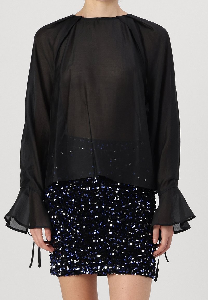 Blouse noire transparente à manches longues avec encolure froncée et poignets évasés ; associée à une jupe courte en sequins bleu marine, alternant des sequins bleus et argentés.
