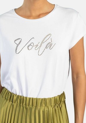 Camisa blanca de manga corta con la palabra «Volfa» escrita con pequeñas piedras marrones y negras, llevada encerrada en una falda plisada de talle alto verde oliva.
