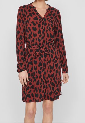 Woman wearing a rust-red button-up dress with black irregular leopard spots, long sleeves, a waist tie, and a chest pocket.