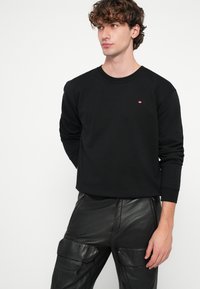 Čierna mikina s dlhým rukávom, okrúhlym výstrihom a malým červeným logom. Kombinovaná s lesklými čiernymi koženými cargo nohavicami so stranovými vreckami.