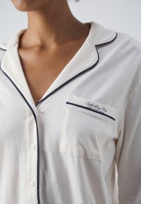 Pyjamashirt i ljusgräddfärgad tyg, med marinblå piping, en bröstficka med branding och knapplåsfunktioner. Mjuk textur och avslappnad passform.