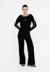 Set de lounge din tricot negru, care include un top cu mâneci lungi, dungi albe și logo, asortat cu pantaloni cu croială largă, prezentat pe un fundal neutru.