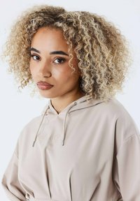 Beige hættetrøje med justerbar snor og broderet logo. Blødt tekstureret stof og afslappet pasform. Modellen har neutral makeup og krøllet hår.