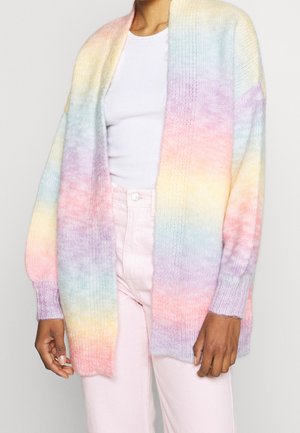 Personne portant un cardigan tricoté aux couleurs pastel arc-en-ciel, un haut blanc côtelé et un pantalon rose clair sur fond uni.