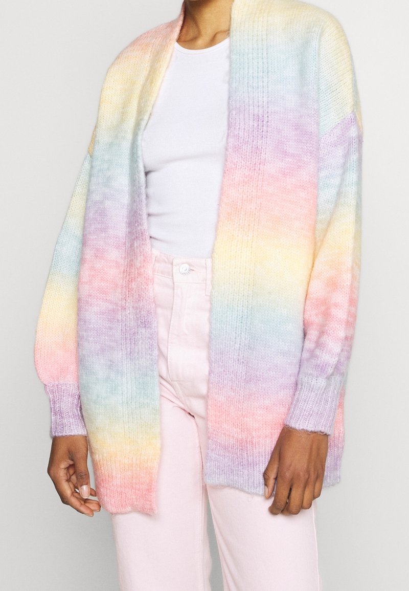 Personne portant un cardigan tricoté aux couleurs pastel arc-en-ciel, un haut blanc côtelé et un pantalon rose clair sur fond uni.