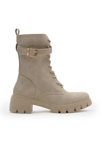 Botín beige con cordones, suela de tracción gruesa, tacón de altura media, correa con hebilla dorada en la parte superior y tirador en la parte posterior.