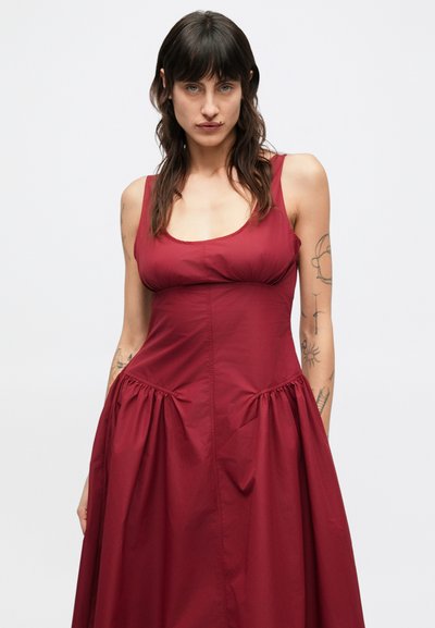 Βουργουνδί φόρεμα χωρίς μανίκια με εφαρμοστό μπούστο και φουσκωτή, πλισέ φούστα. Λείο ύφασμα με ελαφρώς γυαλιστερή υφή, σχεδιασμένο για casual εφαρμογή.