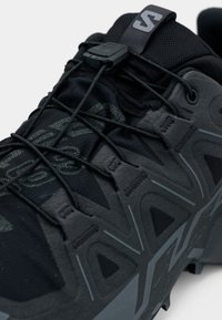 Čierna bežecká trailová obuv s textúrovaným horným dielom z tkaniny, integrovaným šnurovacím systémom, gumovou podrážkou a výrazným geometrickým vzorom na boku.