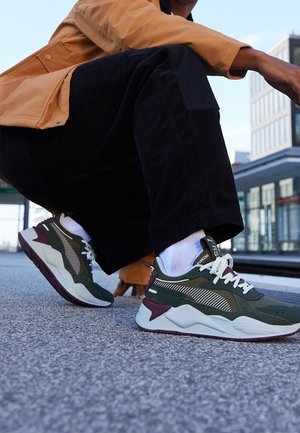 Personne accroupie sur le trottoir portant des baskets de sport vertes et blanches avec un pantalon noir et une veste beige près d'un immeuble moderne.