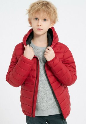 Veste matelassée rouge avec capuche, présentant un motif matelassé diagonal, une fermeture éclair à l'avant et une doublure noire, portée sur une chemise grise.