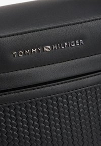 Borsa di pelle nera con trama intrecciata, caratterizzata da un logo "TOMMY HILFIGER" in argento. Le sezioni lisce contrastano con la base a motivo. Dettaglio con cerniera visibile.