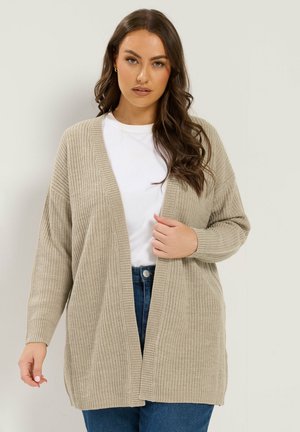 Kvinde med langt bølget hår iført en beige strikket cardigan over en hvid skjorte og blå jeans, poserer mod en ensfarvet lys baggrund.