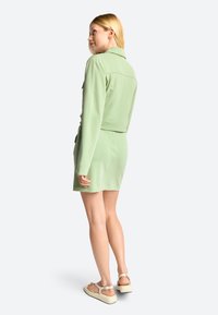Green cropped jacket paired with a matching dress, showcasing smooth fabric, tailored design, and sandals with a neutral color sole.