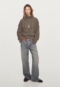 Donna in piedi con le mani in tasca, che indossa un maglione a collo alto marrone, jeans larghi blu chiaro, scarpe scure e una collana con pendente.