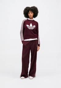 Burgundi kötött pulóver fehér Adidas logóval, csíkos ujjak; hozzá illő bordó bővülő nadrág; mindkettő puha textúrával rendelkezik.