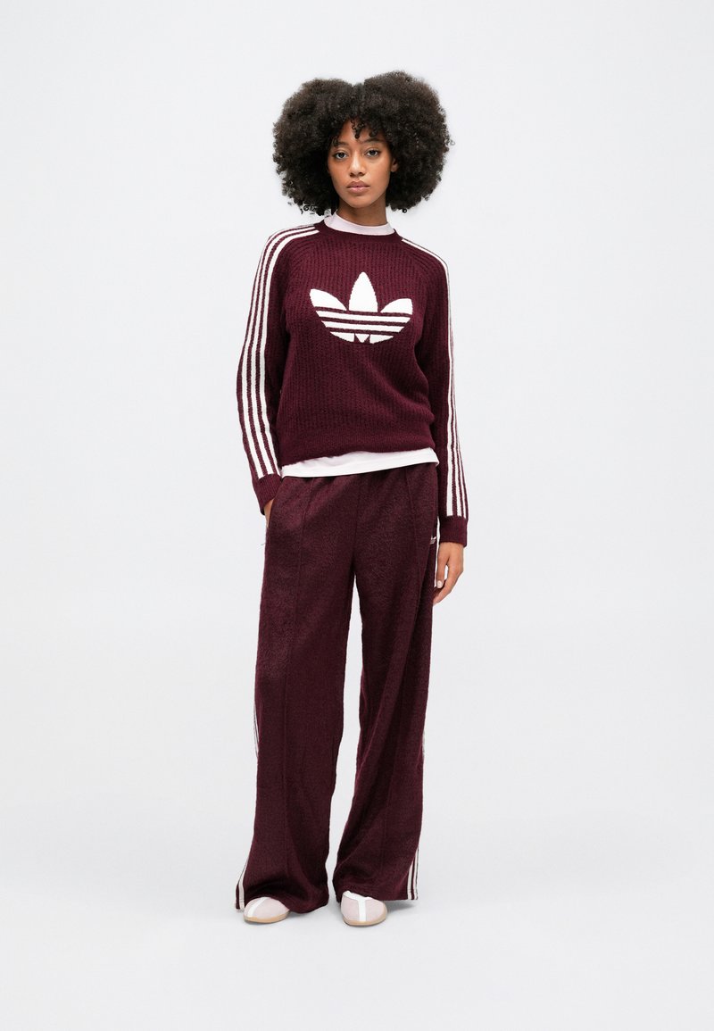 Burgunderfarbener Strickpullover mit weißem Adidas-Logo und gestreiften Ärmeln; dazu passende weinrote weit geschnittene Hose; beide haben eine weiche Textur.