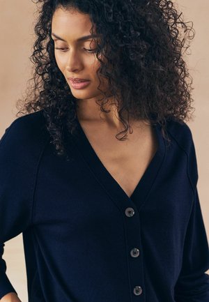 Cardigan bleu marine avec un décolleté en V profond, comportant cinq boutons sombres et un tissu doux et texturé. Les manches ont une coupe décontractée et un design raglan.