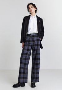 Schwarzer Blazer mit silbernem Akzent, weißes plissiertes Hemd und karierte weite Hosen in Marineblau und Grau. Schwarze Schuhe mit dicken Sohlen.