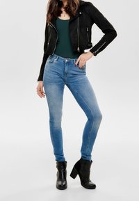 Zwarte suède cropped jack, groene tanktop, lichtblauwe skinny jeans en zwarte enkellaarsjes met een blokhak, tegen een neutrale achtergrond.