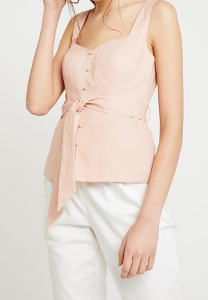 Vrouw draagt een mouwloze lichtroze top met knopen en een tailleband die is gestrikt, gecombineerd met een witte broek, tegen een effen achtergrond.