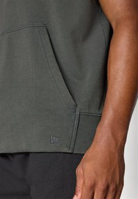 Sudadera verde oscuro con un bolsillo tipo canguro, ribete acanalado en el dobladillo y un logo sutil en la parte inferior izquierda. Se puede apreciar la textura suave de la tela.