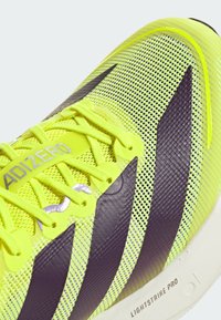 Scarpa da atletica di un brillante colore giallo, con tomaia in rete, strisce di un viola scuro e dettagli grigio chiaro. Presenta una superficie testurizzata e lacci visibili.