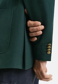 Blazer vert texturé avec une poche plaquée et des accents de boutons dorés sur les manches. Gros plan montrant une main tenant le tissu.