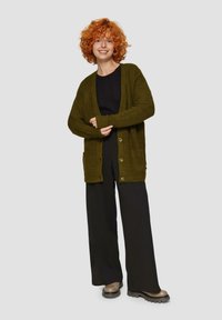 Olivgrüner, gestrickter Cardiagan mit Fronttaschen, fünf Knöpfen und lässigem Schnitt, kombiniert mit einem schwarzen Oberteil und weiten schwarzen Hosen.