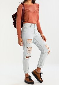 Haut à manches longues couleur rouille avec col à volants, associé à un jean bleu clair déchiré. Chaussures marron à lacets avec semelles noires. Sac à dos rouge.