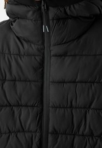 Czarna ocieplana kurtka puchowa z pikowanym wzorem, wysokim kołnierzem oraz przednim zamkiem błyskawicznym z regulacją sznurkową przy kapturze.