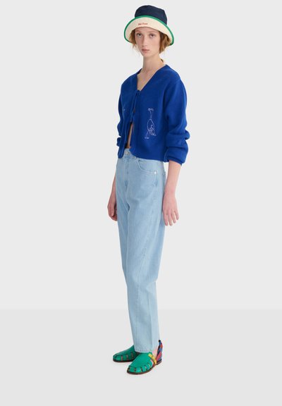 Jeune personne portant un cardigan bleu avec une broderie d'oiseau, un jean bleu clair, des sandales vertes et un bob bicolore, debout de profil.