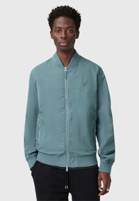 Giacca bomber in un leggero teal con zip frontale, tasche laterali, colletto e polsini a coste. Presenta un piccolo logo sul lato sinistro del petto. Tessuto morbido.