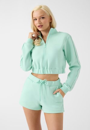Femme aux longs cheveux blonds portant une veste courte zippée vert menthe et un short à cordon assorti, debout sur un fond neutre.