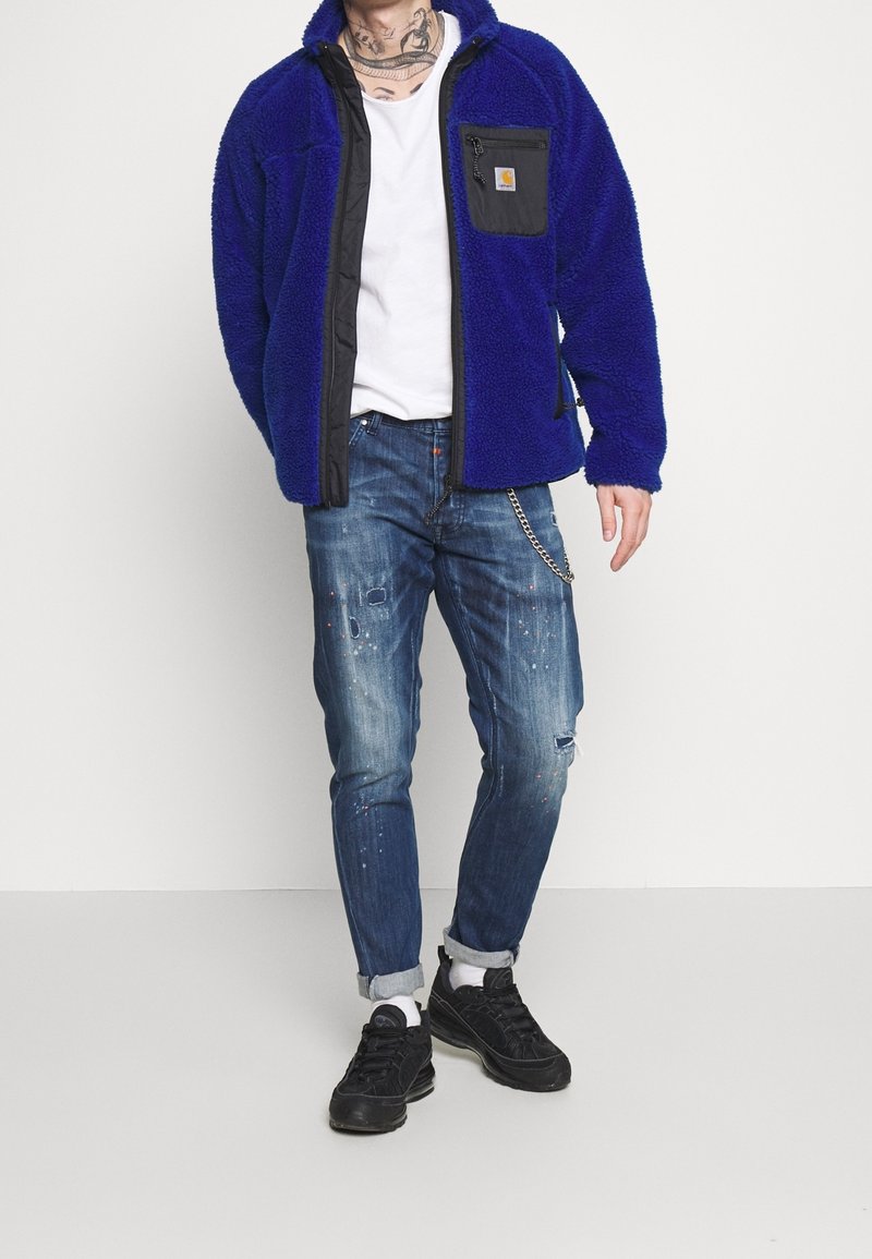Modrá fleecová bunda s černými detaily, zipem a náprsní kapsou, spárovaná s obnošenými modrými džíny a černými teniskami.