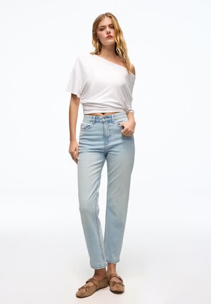 Fiatal nő laza, fehér, egypántos felsőt, világoskék magasderekú farmert és barna, felhúzható cipőt visel, és fehér háttér előtt áll.