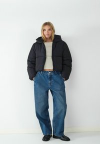 Czarna kurtka puchowa z kapturem, pikowanymi sekcjami i zamkiem błyskawicznym, w połączeniu z luźnymi niebieskimi dżinsami i czarnymi butami.