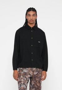 Camisa preta de manga longa com botões à frente, um bolso no peito com o logo "OBEY" e um corte descontraído. Combinada com calças castanhas padronizadas.