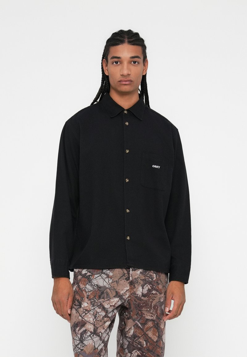 Camisa preta de manga longa com botões à frente, um bolso no peito com o logo "OBEY" e um corte descontraído. Combinada com calças castanhas padronizadas.