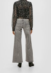 Person, der bærer højtaljede grå brede jeans og en gennemsigtig sort blomstret bluse med lange ærmer, står foran en ensfarvet baggrund.