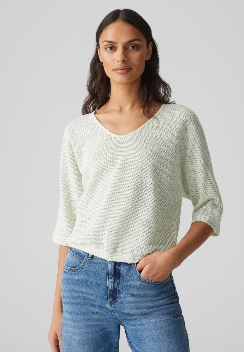 Helles grünes gestricktes Oberteil mit V-Ausschnitt, dreiviertellangen Ärmeln und einem strukturierten Muster; kombiniert mit blauen Jeans.