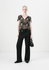 Donna in piedi con pantaloni neri a gamba larga con bottoni dorati, top corto a maniche corte con scollo a V a fantasia, scarpe nere in vernice, mentre tiene una borsa nera.