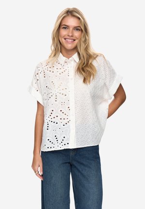 Lächelnde Frau mit langen blonden Haaren, die eine kurzärmelige weiße Bluse mit Ösenmuster und blaue Jeans trägt und mit einer Hand auf der Hüfte steht.